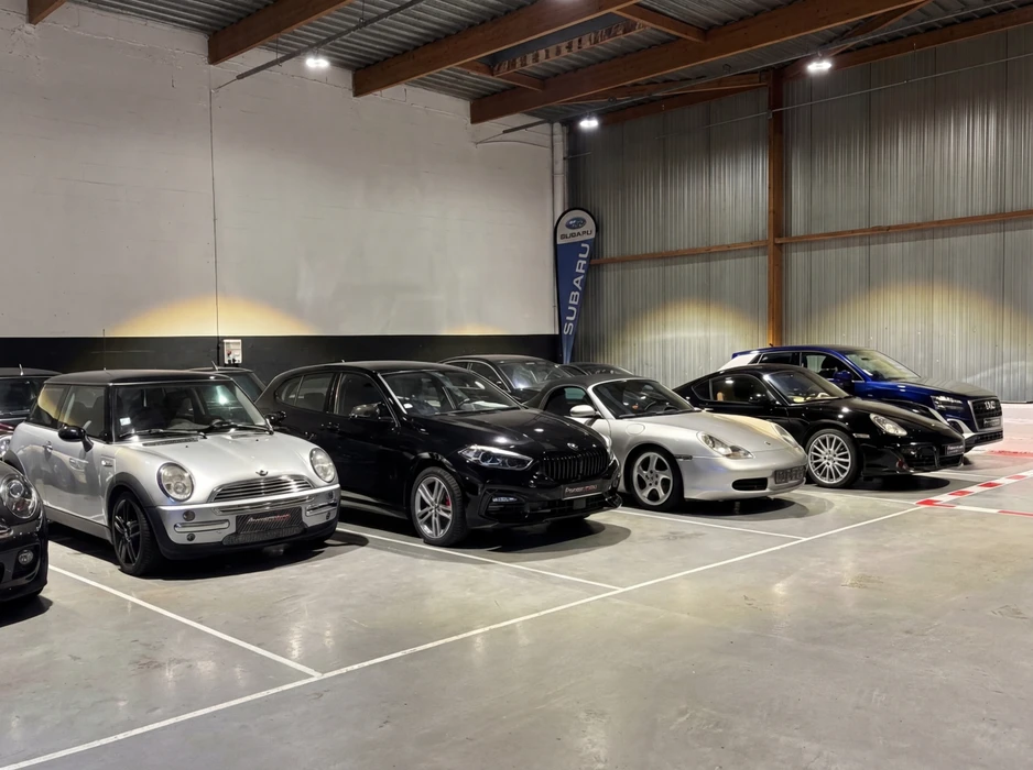 Showroom automobile intérieur avec 5 véhicules alignés : une Mini Cooper argentée, un BMW X6 noir, une Porsche 996 Carrera argentée, et une Mercedes noire/bleue