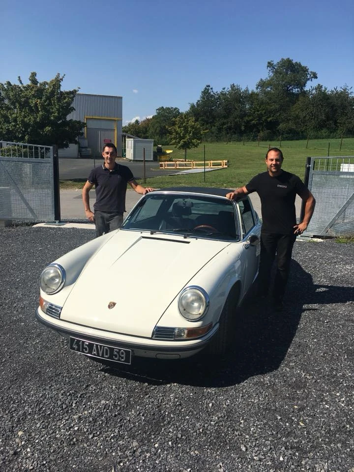 Photographie professionnelle en intérieur d'un atelier automobile spécialisé montrant un mécanicien ou technicien automobile effectuant une inspection détaillée d'une voiture haut de gamme (Porsche, Mercedes ou BMW par exemple). La scène doit montrer clairement des outils de diagnostic professionnels, le véhicule sous bon éclairage LED, des équipements d'atelier moderne, et une atmosphère de précision technique. Le véhicule doit être visible entièrement ou partiellement, avec focus sur le professionnalisme de l'inspection en cours. Ambiance sérieuse, éclairage blanc froid professionnel.