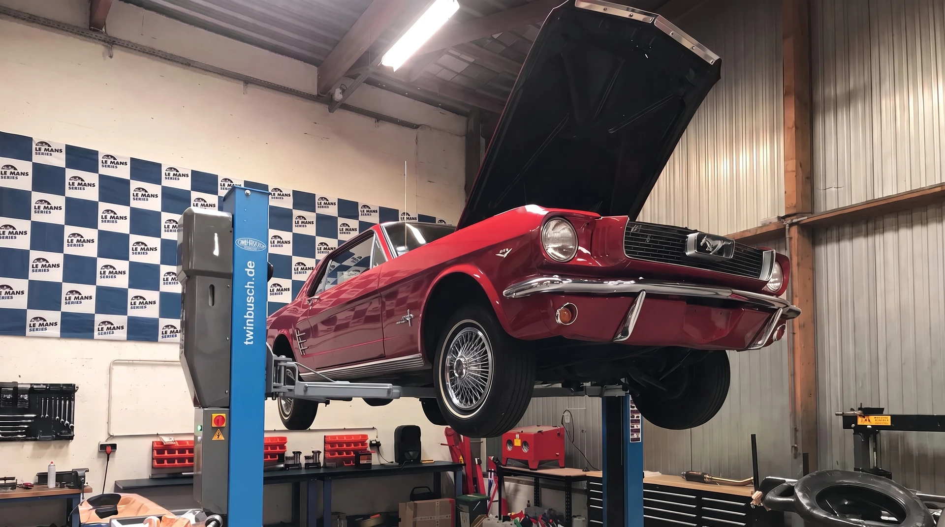Photographie d'intérieur d'un atelier automobile professionnel spécialisé, montrant une voiture de collection prestigieuse (Ferrari ou Porsche classique) stationnée sur un pont élévateur hydraulique à deux colonnes bleu. Le véhicule est vu de trois-quarts avant, révélant des détails d'excellente restauration : carrosserie impeccable, jantes alliage brillantes, phares chromés d'époque. En arrière-plan, un mécanicien en tenue professionnelle travaille, entouré d'outillage spécialisé (servante d'atelier, extracteurs, équipement de diagnostic). L'atelier présente une structure métallique apparente, sol époxy, éclairage LED industriel. Une enseigne Presteam Automobiles et une bannière Le Mans sont visibles. L'ambiance générale doit transmettre l'expertise, la précision et la passion pour les véhicules de collection.